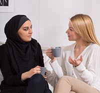A woman wearing a Hijab speaking to another woman over mugs of tea
