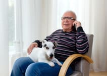 An older man sitting in a chair with his dog. The man is using his mobile phone.