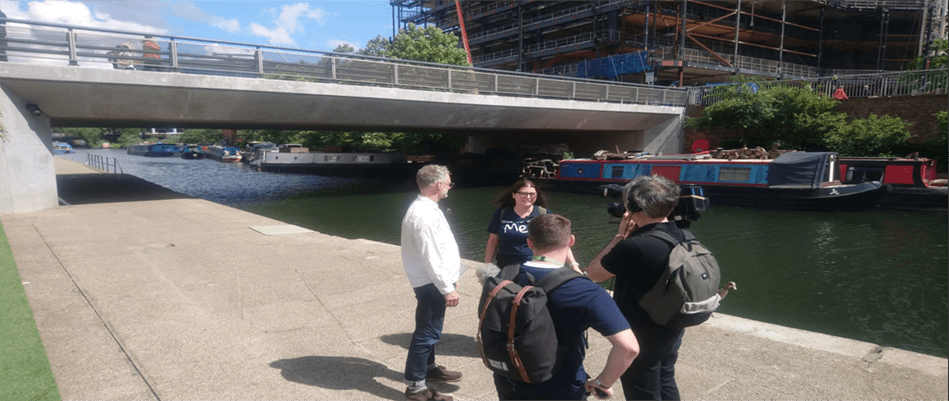 Helen Moulinos is speaking to a male member of the public by a river. There is a bridge and a canal boat in the background. They are being filmed by a cameraman  for our Human Rights Voxpops series.